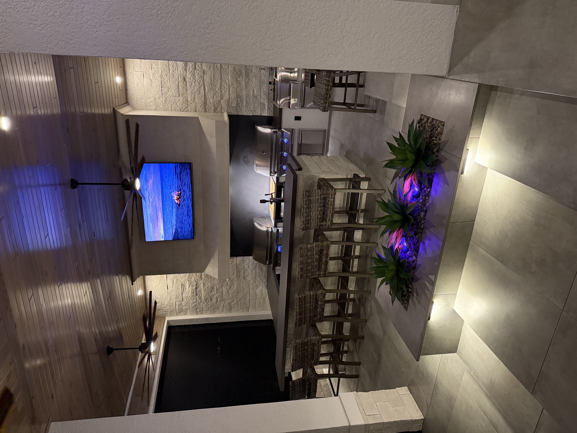 The finished outdoor living space lit at night with bar stools and TV wall